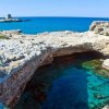 Acque limpide sotto una scogliera rocciosa, con torre lontana e mare blu.