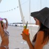 Donna con cappello beve cocktail in spiaggia, un'altra persona sullo sfondo con telo viola.