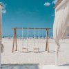 Spiaggia sabbiosa con altalena in legno tra tende bianche e mare azzurro sullo sfondo.
