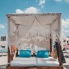 Letto da spiaggia con tenda intrecciata, due cuscini blu, su una spiaggia sabbiosa.
