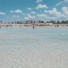 Spiaggia con acqua trasparente e edifici sullo sfondo, cielo azzurro con nuvole.