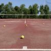 Campo da tennis rosso con palline gialle sparse, circondato da alberi e rete.