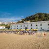 Una spiaggia sabbiosa con lettini e ombrelloni davanti a un grande edificio bianco e colline verdi.